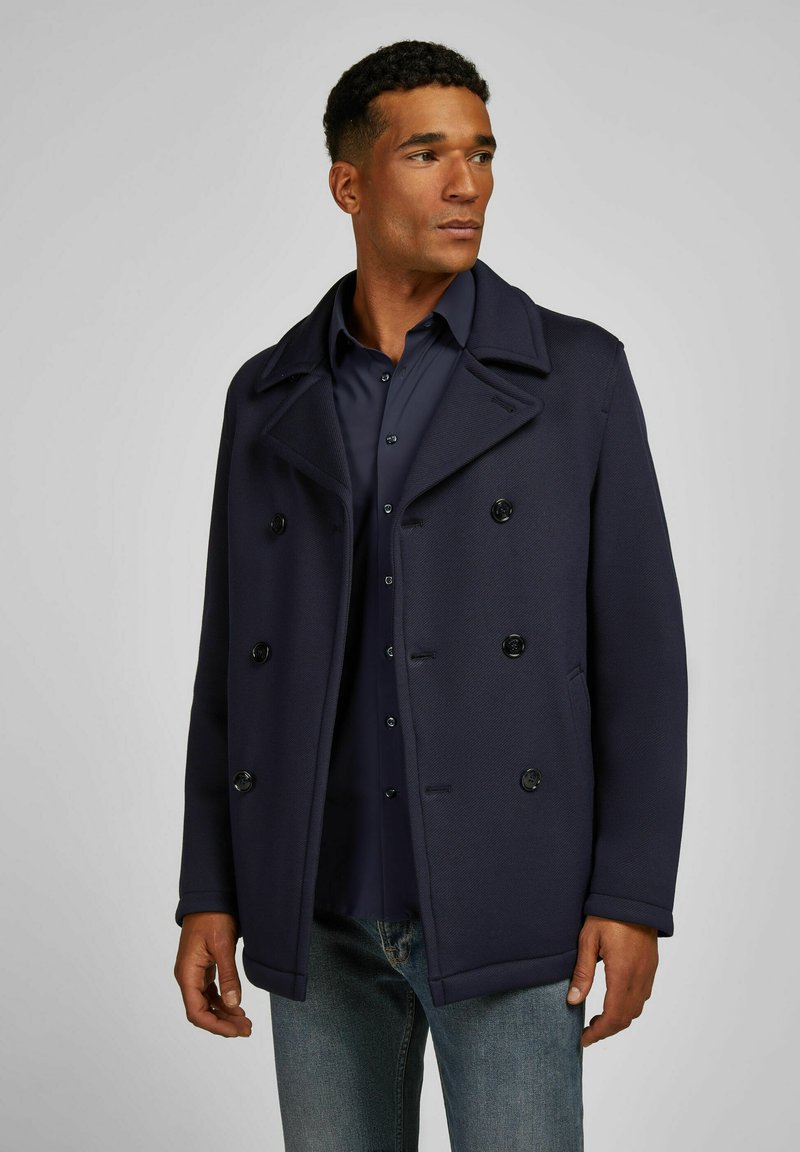 Marineblauer, doppelt geknöpfter Mantel mit großen schwarzen Knöpfen, spitzem Kragen und strukturiertem Stoff, getragen über einem dunklen Hemd und Jeans.