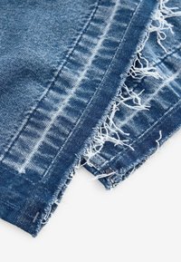 Gros plan sur les bords effilochés de l'ourlet d'un jean en denim bleu, avec des fils blancs visibles et des détails de couture décolorée.