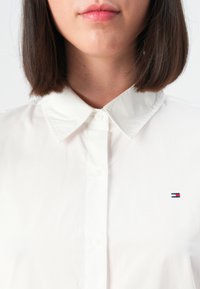 Femme aux cheveux bruns lisses portant une chemise blanche boutonnée avec col et un petit logo rectangulaire rouge, blanc et bleu sur la poitrine.