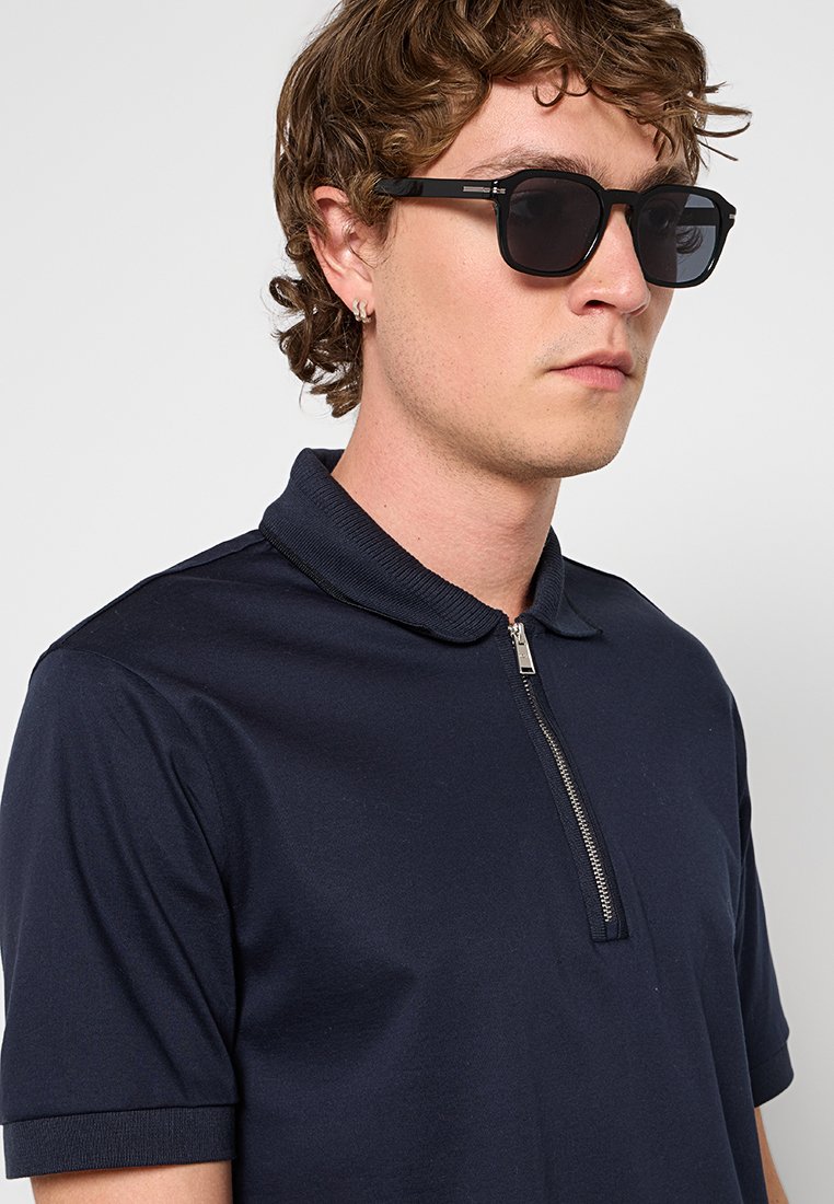 Polo shirt marine avec un col zippé, des manches courtes et une texture de tissu lisse. Porté avec des lunettes de soleil noires, présentant une monture carrée moderne.