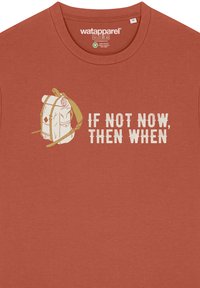 Koperkleurig katoenen T-shirt met een grafisch ontwerp van een rugzak en de tekst "ALS NU NIET, WANNEER DAN" in het wit. Ronde halslijn, korte mouwen.