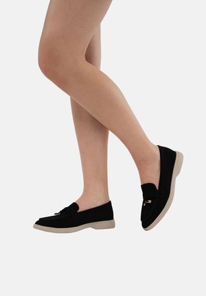Mocassins en daim noir avec une semelle beige plate, ornés d'un accent en nœud et de petits éléments en or, présentés sur une jambe croisée.