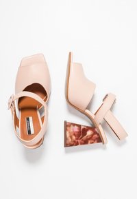 Pár světle růžových sandálů na podpatku s otevřenou špičkou, silnými blokovými podpatky s mramorovým vzorem a kotníkovými páskami na bílém pozadí.