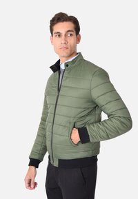 Giacca imbottita verde con colletto alto, zip frontale e polsini a coste neri. Presenta tasche laterali e una texture liscia e lucida.