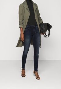 Olijfgroene trenchcoat over een zwarte coltrui, gecombineerd met donkere spijkerbroek en zwarte sandalen met hoge hakken, met een zwarte tas in de hand.