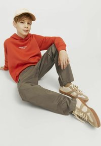 Röd huvtröja, olivgröna cargobyxor och beige sneakers med accenter. Ungdom som sitter och poserar avslappnat mot en vit bakgrund.
