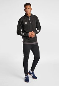 Schwarze athletische Jacke mit Viertel-Reißverschluss und grauen Akzenten, lange Ärmel mit reflektierenden Mustern, dazu slim-fit schwarze Sporthose. Blaue Sportschuhe.