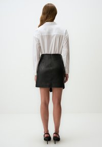 Mini-jupe en cuir noire au design ajusté, fermeture à glissière dans le dos et détails de plis légers. Associée à une chemise blanche à manches longues.