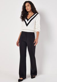 Maglione bianco con scollo a V nero e motivo a zigzag abbinato a pantaloni neri a vita alta. L'outfit presenta una vestibilità sartoriale e una texture liscia.