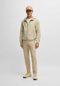 Light beige windbreaker with hood, front zipper, and two side pockets. Paired with beige pants and cream sneakers. Beige cap with logo.