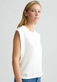 Valge ilma varrukateta särk, mis on valmistatud pehmest kangast, laia lõikega, ümmarguse kaelusega ja dekoratiivsete detailidega allservas.