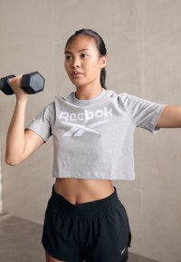 Grå cropped t-skjorte med hvit "Reebok" logo, kombinert med svarte treningsshorts. Personen løfter en sekskantet svart manual.
