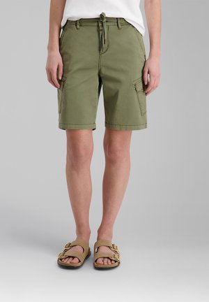 Person trägt olivgrüne knielange Cargoshorts, ein weißes Hemd und beige Sandalen mit goldenen Schnallen, steht vor einem einfarbigen grauen Hintergrund.