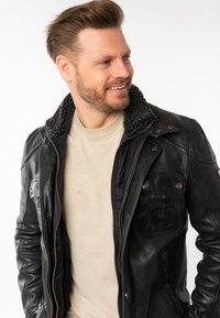 Zwarte leren jas met een gestructureerde, zwarte schapenvachtkraag, voorzakken en zilverkleurige hardware. Draagt over een lichtbeige shirt.
