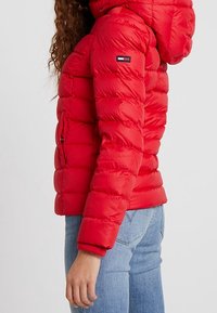 Kvinde iført rød, afdæmpet pufferjakke med hætte og lyseblå jeans, stående i profil mod en ensfarvet baggrund.