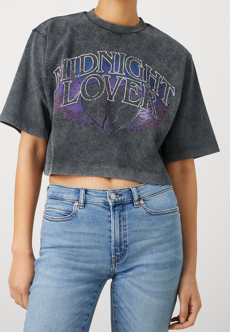 Camiseta negra corta con el texto "MIDNIGHT LOVER" y un gráfico de alas en púrpura. Combinada con jeans ajustados de color azul claro, con un diseño clásico de cinco bolsillos.
