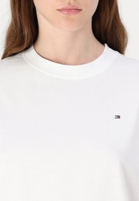 Hvid bomulds t-shirt med rund halsudskæring, der har et lille broderet logo i rød, blå og hvid på venstre bryst. Glat tekstur.