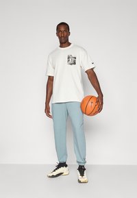 T-shirt blanche à manches courtes avec une poche graphique noir et blanc, associée à un pantalon de survêtement bleu clair et des baskets noir et blanc, tenant un ballon de basket.
