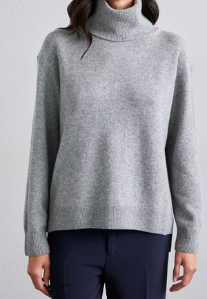 Femme portant un pull à col roulé en tricot gris clair à manches longues et un pantalon bleu marine foncé sur un fond uni.