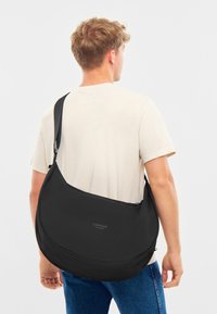Černá ramenní taška s zakřiveným designem, vyrobená z hladkého materiálu, s logem a nastavitelným popruhem, nošena přes tělo.