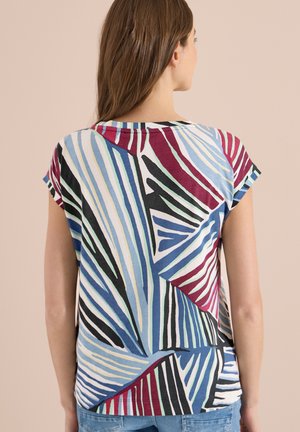 Vrouw gezien van achteren die een top met korte mouwen draagt met geometrische multicolor strepen in blauw, zwart, rood en wit.