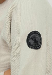 Beige stickad ärm med en svart cirkulär patch som visar en jordglob och texten "Jack & Jones // Global Community // Core // Est. 1990".