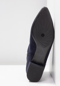 Chaussure en daim bleu marine à bout pointu avec semelle en caoutchouc noire indiquant la taille 4 1/2, positionnée verticalement contre une surface blanche.