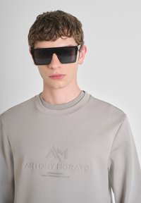 Sudadera gris claro con detalles de logo en relieve; modelo que lleva gafas de sol negras angulares grandes, pelo corto y texturizado oscuro; fondo neutro.