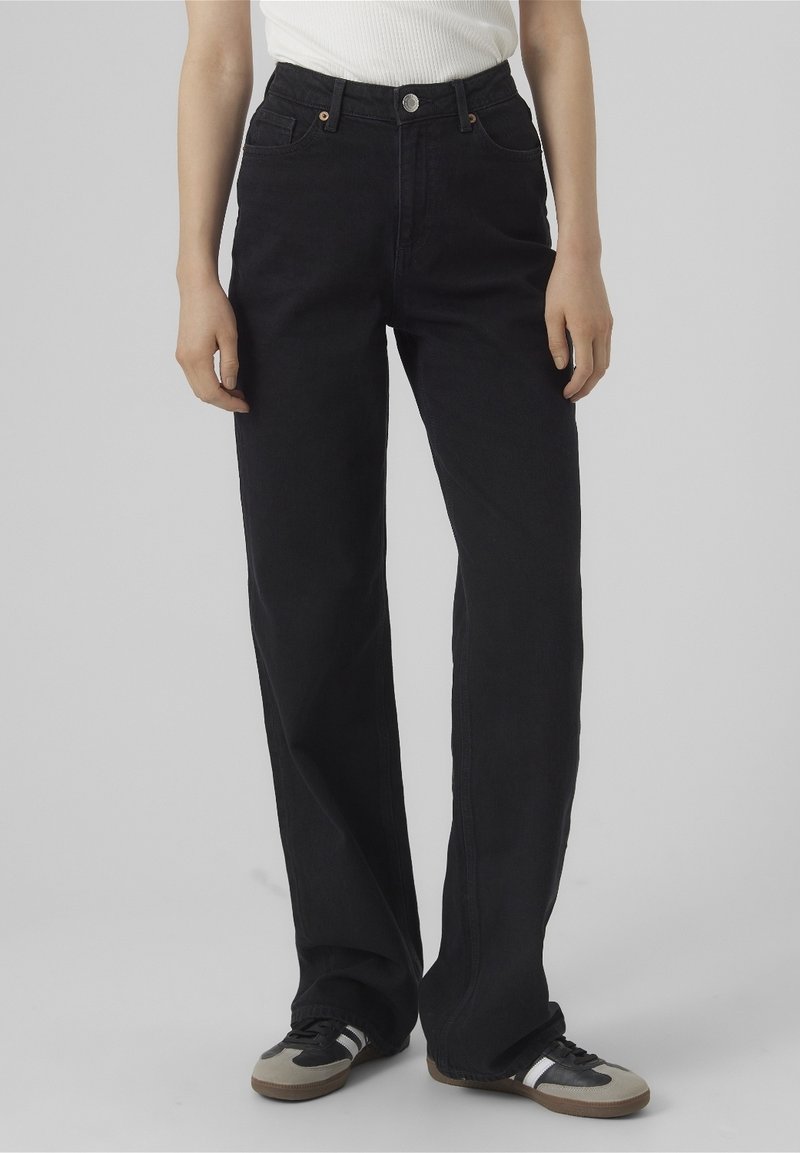 Personne portant un jean noir taille haute à jambes droites associé à des baskets noires et beiges sur un fond neutre.