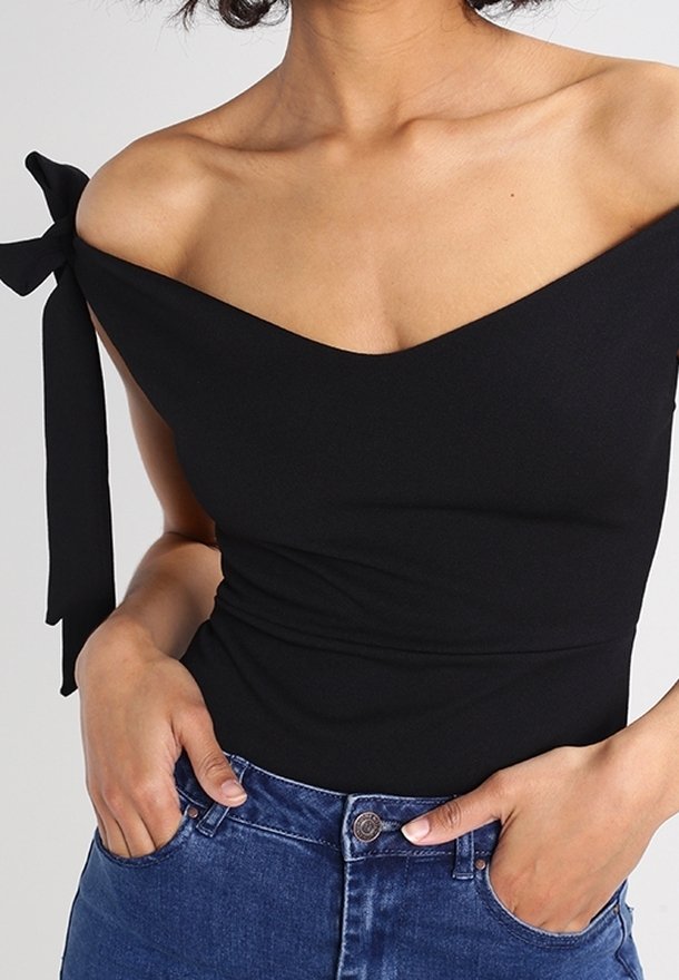 Femme portant un haut noir à épaules dénudées avec un nœud sur l'épaule gauche, assorti à un jean bleu taille haute, les mains dans les poches.