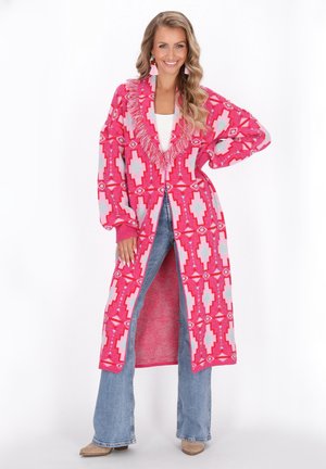 Roze cardigan met geometrische patronen in blauw en wit, voorzien van een franjeshals en brede mouwen, gedragen over lichte denim flairs.