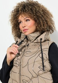 Beige Steppweste mit schwarzen Lin Zeichnungen, hohem Kragen, Reißverschluss und verstellbaren Kordeln. Tragend über einem schwarzen Langarmoberteil.