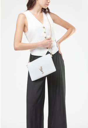 Borsa a tracolla bianca con texture e logo dorato, dotata di tracolla a catena. Il modello indossa un gilet bianco e pantaloni larghi neri.