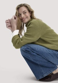 Groene gebreide trui met een hoge hals, gecombineerd met losse blauwe spijkerbroeken en donkere glanzende schoenen. Het model is in een hurkpositie.