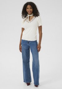 Cremig geripptes Kurzarmoberteil mit gewelltem Saum und Spitzen-Neckline, kombiniert mit blauen Schlagjeans und kastanienbraunen Absatzschuhen. Texturiertes Schal-Accessoire.
