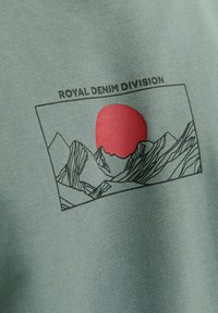 Zöld anyagú póló, amelyen fekete grafikai ábrázolás látható hegyekről és egy piros napról, a rajz felett pedig a "ROYAL DENIM DIVISION" felirat olvasható.