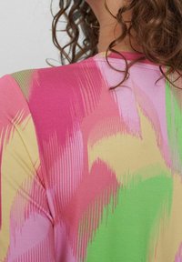 Une personne aux cheveux bouclés portant une chemise multicolore avec des motifs de coups de pinceau abstraits en rose, vert, jaune et rouge.