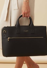 Sac à main en cuir noir avec une finition texturée, doté de deux poignées supérieures, de quincaillerie dorée et d'une poche frontale zippée. Détail du logo doré visible.