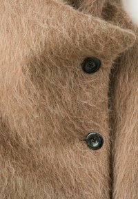 Palton bej călduros din blană moale, cu nasturi negri mari și guler larg cu revere, evidențiind țesătura moale și texturată și cusăturile detaliate ale nasturilor.