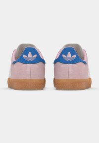 Par de ténis em camurça rosa com abas azuis no calcanhar, apresentando um logótipo trefoil rosa da Adidas e solas de borracha de gengibre, vistos pelo lado de trás.