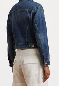 Dark blue denim jacket with front pockets, a collared neck, and cuffed long sleeves, shown from the back, paired with light-colored trousers.