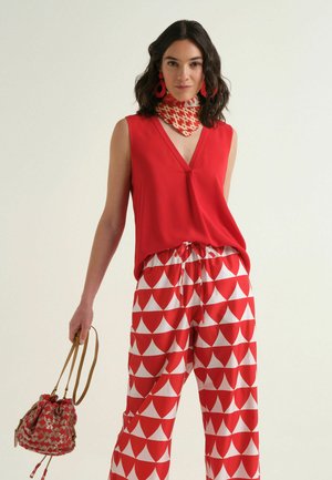 Femme en haut rouge sans manches et pantalon large à motifs rouge et blanc tient un sac assorti avec motif rouge, porte des boucles d’oreilles rouges et un foulard autour du cou.