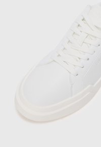 Adidași din piele albă cu vârful rotund, șireturi plate și o talpă texturată. Prezentă un branding minim și detalii de cusătură discrete.