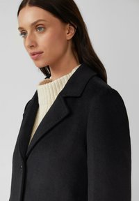 Schwarzer Wollmantel mit eingeschlagenem Kragen, einer Knopfverschluss und darunter ein strukturierter cremefarbener Pullover mit geripptem Muster.