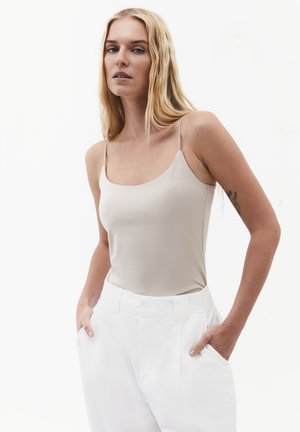 Blonde vrouw met lang haar, die een beige mouwloze top en een witte broek draagt, staat met de handen in de zakken tegen een witte achtergrond.