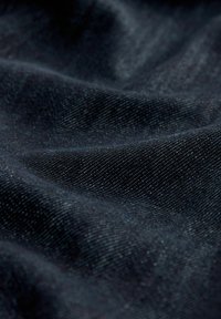 Tamno plavi denimski materijal s suptilnom teksturom, koji ima glatku površinu i blagi sjaj, prikazuje preklopljene i nabore.