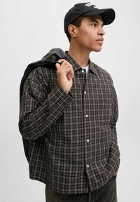 Jonge man in een geruite shirt en zwarte pet, met een leren jacket over zijn schouder, die bedachtzaam opzij kijkt.