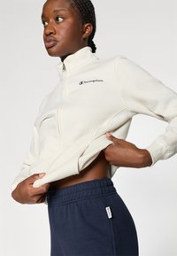 Sudadera cremosa con cremallera y logo "Champion" en negro, combinada con pantalones de chándal azul marino que tienen una cinturilla elástica y una etiqueta con el logo.