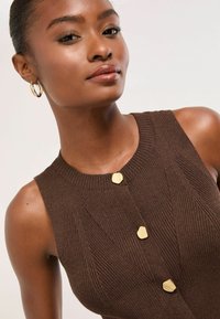 Brown ribbed sweater with a round neckline, featuring three gold button accents and a fitted silhouette. Smooth texture with subtle patterning.