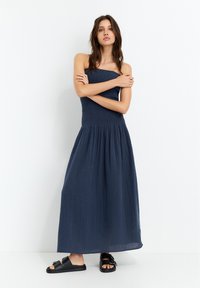 Navy-blaues Maxikleid mit gerafftem Oberteil, fließendem, plissiertem Rock und ärmellosem Design, kombiniert mit schwarzen Sandalen mit dekorativen Schnallen.